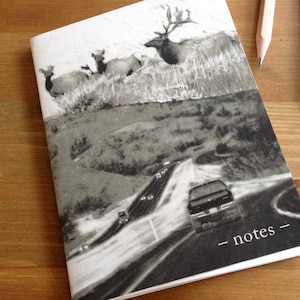May include: A black and white photo of a notebook with a scenic image of a highway with cars driving on it. The image also includes a group of elk in the background. The notebook has the word "notes" written on the front cover.