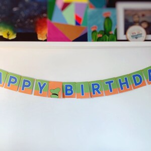 May include: A green and orange banner with blue letters that reads "Happy Birthday". The banner has a small green hat with a yellow brim on the "B".