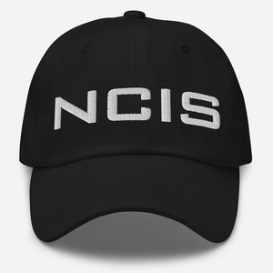 Può includere: Cappellino da baseball nero con il testo "NCIS" ricamato in bianco.