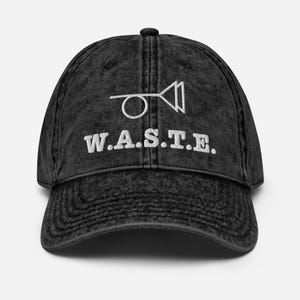 Peut inclure: Casquette de baseball noire délavée avec une visière incurvée. Le chapeau présente un texte brodé blanc et un graphique d'une corne et de deux triangles. Le texte se lit "W.A.S.T.E." dans une police sans-serif en gras. La casquette a un style vintage classique.