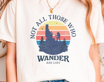 Op de afbeelding: Een crèmekleurig T-shirt met korte mouwen en een grafische print van een wandelaar op een bergtop tegen een zonsondergang. De tekst NOT ALL THOSE WHO WANDER ARE LOST staat in een cirkel rond de centrale bergafbeelding.
