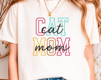 Puede incluir: Camiseta color crema con el texto "Cat Mom" en letras de colores. La parte superior se combina con vaqueros azules. La camiseta es una prenda informal para el día a día.