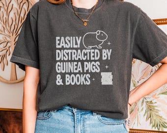 May include: Dark gray t-shirt with the text "EASILY DISTRACTED BY GUINEA PIGS & BOOKS" in white. The shirt features a guinea pig graphic. The model is wearing light blue jeans.