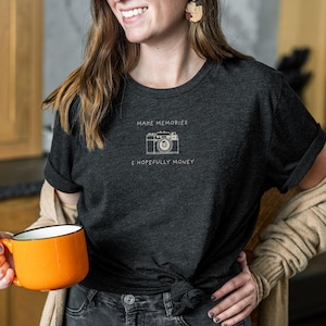 Op de afbeelding: Een donkergrijs T-shirt met een witte afbeelding van een camera en de tekst "Make Memories & Hopefully Money".
