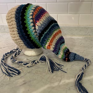 May include: A colorful, handmade crochet hat with a long, pointed tail and braided tassels. The hat features a mix of cream, blue, green, orange, and purple yarns. The tassels are blue and cream. The hat is displayed on a white foam head.