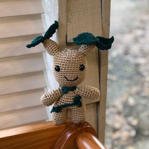 May include: A tan crochet doll with black eyes, a smiling mouth, and dark green leaf-like accents on its head and chest. The handmade doll has a whimsical design, perfect for a child's toy or a unique decorative item.
