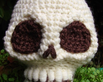 May include: A white crocheted skull with brown yarn eyes. The skull is sitting on a bed of green grass.