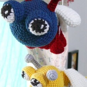 May include: Two crocheted toy airplanes hanging from a ceiling. The first airplane is blue, red, and white. The second airplane is yellow, white, and gray.