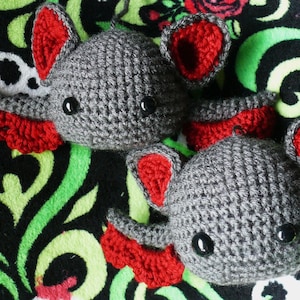 May include: Two gray crocheted bats with red felt wings and black eyes. The bats are sitting on a black and green patterned fabric.