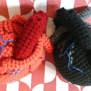 May include: Two crocheted hearts, one red and one black, with blue stitching representing veins. The hearts are on a red and white patterned background.