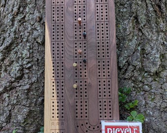 May include: A wooden cribbage board with a dark finish, featuring drilled holes and metal pegs. A deck of Bicycle playing cards rests beside the board. The board is approximately 15 inches long.