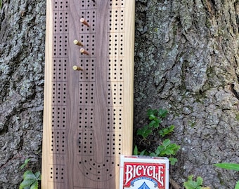 May include: A wooden cribbage board with a dark walnut center and light wood edges. The board has drilled holes for pegs and is accompanied by a deck of Bicycle playing cards. The pegs are black and copper-colored.