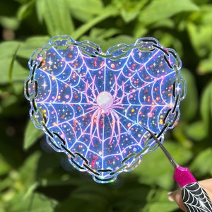 May include: A heart-shaped sticker with a pink spider in a web design. The web is filled with glitter and the sticker has a chain link border.