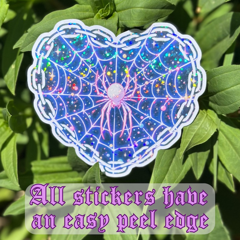 May include: A heart-shaped sticker with a glittery, iridescent finish. The sticker features a pink spider in a web with a pearl-like body. The text "All stickers have an easy peel edge" is printed below the sticker.