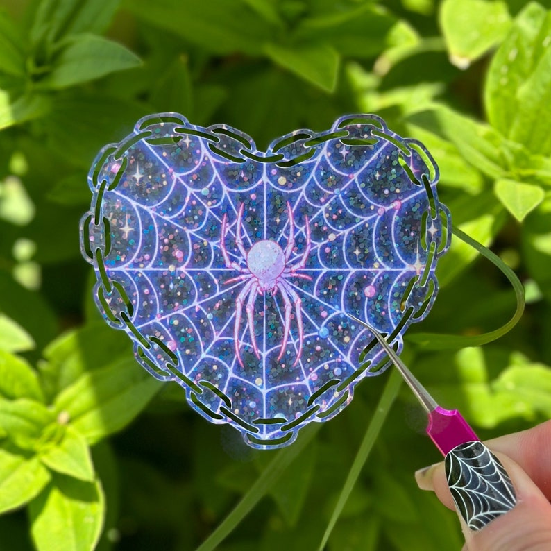 May include: A heart-shaped sticker with a glittery purple and blue spiderweb design. A pink spider is in the center of the web. The sticker has a chain-link border.
