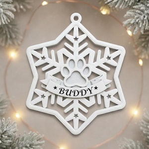 Op de afbeelding: Wit sneeuwvlokvormig ornament met een pootafdruk en de naam "BUDDY" op een banner. Het ornament heeft een sterontwerp en is waarschijnlijk voor een huisdier. Het ornament hangt aan een lus aan de bovenkant.