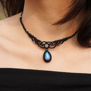 May include: A black cord necklace with a silver and black intricate design. A teardrop-shaped blue gemstone hangs from the centre of the necklace.