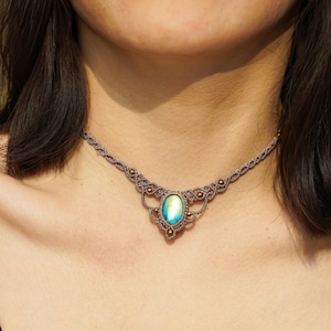 Puede incluir: Un delicado collar gargantilla con un cordón marrón claro y un gran colgante de piedra preciosa iridiscente de forma ovalada. El colgante está rodeado de pequeñas cuentas doradas.