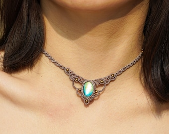 Puede incluir: Un delicado collar gargantilla con un cordón marrón claro y un gran colgante de piedra preciosa iridiscente de forma ovalada. El colgante está rodeado de pequeñas cuentas doradas.