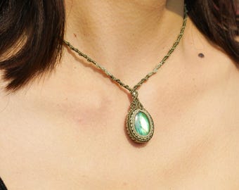 Puede incluir: Un colgante de piedra preciosa verde e iridiscente en un collar de cordón de macramé. El colgante tiene forma ovalada y tiene un diseño tejido alrededor de la piedra.