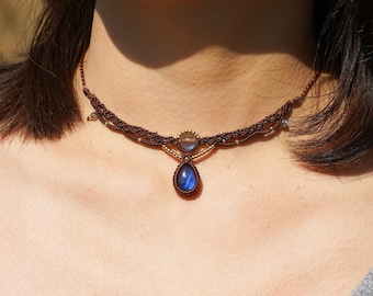 Puede incluir: Un collar gargantilla de macramé marrón con un colgante de piedra preciosa azul en forma de lágrima y una piedra preciosa redonda más pequeña.