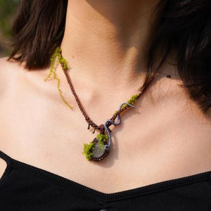 May include: A brown cord necklace with a green moss and gray stone pendant. The necklace is made with a woven cord and features a unique design with a small gray stone in the center of the pendant.