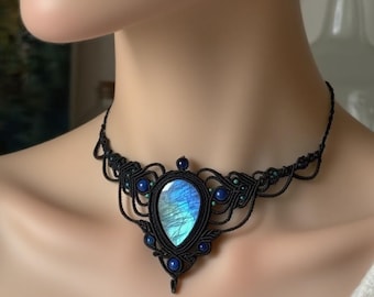 Puede incluir: Un collar de macramé negro con una gran piedra preciosa azul iridiscente en forma de pera. El collar está adornado con cuentas azules más pequeñas y un intrincado trabajo de nudos, creando un diseño elegante y artesanal.