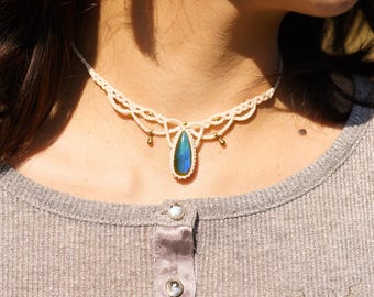 May include: A white macrame choker necklace with a teardrop-shaped blue labradorite gemstone pendant. The pendant has a gold setting and is accented with small gold beads.