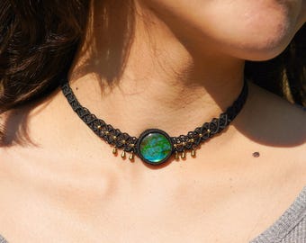 Puede incluir: Un collar gargantilla negro con un colgante redondo de piedra preciosa verde y azul iridiscente. El collar tiene cuentas y borlas doradas.