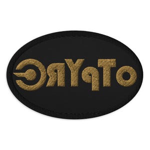May include: Black oval patch with gold embroidered text that reads "ORYATO". The patch has a black border and the text is in a stylized font. The patch is likely for clothing or accessories.