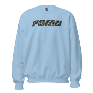 Puede incluir: Una sudadera de color azul claro con la palabra "FOMO" impresa en la parte delantera en un patrón de cuadros negros y dorados.