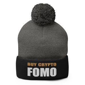 Puede incluir: Un gorro gris y negro con un pompón en la parte superior. El gorro tiene el texto "BUY CRYPTO FOMO" en letras blancas y doradas.