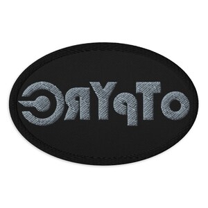 May include: Black oval patch with embroidered gray text that reads "OTYRC". The patch has a black border and the text is in a stylized font. The patch is likely designed to be sewn onto clothing or accessories.