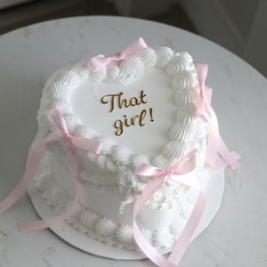 May include: A heart-shaped cake with smooth white frosting, decorated with piped white icing borders and several light pink satin ribbons tied in bows. The top of the cake features the gold-colored text That girl! in a cursive script font.
