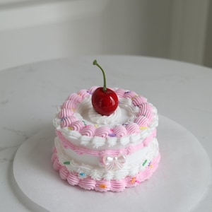 May include: A small, two-tiered cake decorated with pink and white frosting, colorful sprinkles, and a single red cherry on top.