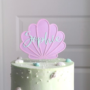 May include: A light green cake decorated with white sprinkles and seashell accents. A pink seashell cake topper with the name "Sophie" in light blue script is placed on top. The cake is on a white surface.