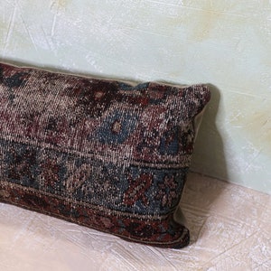 Puede incluir: Una almohada larga y rectangular con una tela estampada en azul oscuro, rojo y crema. La almohada tiene un borde deshilachado y un aspecto desgastado sutil.