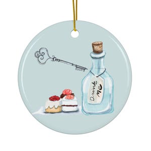 Puede incluir: Adorno redondo de cerámica con fondo azul claro, con una ilustración caprichosa. Se representa una llave, una botella con la etiqueta "Drink me" y dos pequeños pasteles con detalles rojos y blancos. El adorno cuelga de un cordón dorado.