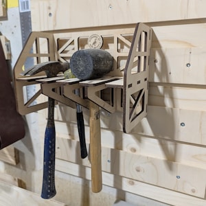 Puede incluir: Un organizador de pared de madera con cuatro ranuras para sujetar martillos y un mazo. El organizador está hecho de madera clara y tiene un diseño simple. Está montado en una pared de madera clara.