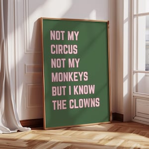 以下が含まれることがあります： 緑色の背景にピンク色の文字で「NOT MY CIRCUS NOT MY MONKEYS BUT I KNOW THE CLOWNS」と書かれたフレーム入りのポスター。 フレームは金色で、ポスターは白い壁と木の床に立てかけられています。