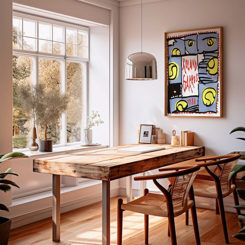 May include: A wooden table with a rustic finish sits in a room with a large window and a poster on the wall. The poster is a colorful illustration of tennis balls and the text "Roland Garros 1988". Two chairs with woven seats are positioned near the table.