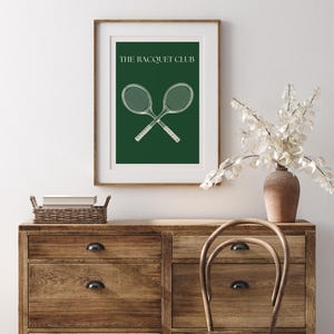 May include: A framed print with a green background and white text that reads "The Racquet Club". Two white tennis rackets are crossed in the center of the print.