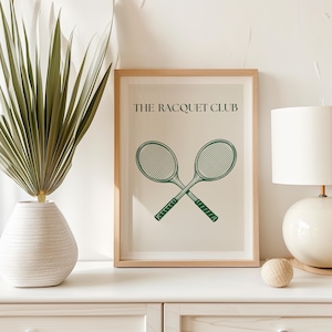 Puede incluir: Una impresi&oacute;n enmarcada con un marco marr&oacute;n claro y un fondo blanco. La impresi&oacute;n presenta una ilustraci&oacute;n verde de dos raquetas de tenis cruzadas y el texto "THE RACQUET CLUB".