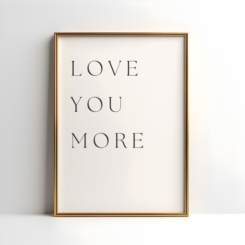 K&ouml;nnte beinhalten: Ein goldfarbener gerahmter Druck mit dem Text "LOVE YOU MORE" in schwarzen Buchstaben auf wei&szlig;em Hintergrund.
