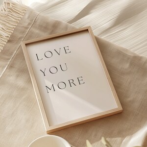 K&ouml;nnte beinhalten: Ein gerahmter Druck mit dem Text "LOVE YOU MORE" in schwarzen Buchstaben auf wei&szlig;em Hintergrund. Der Rahmen ist aus hellem Holz.