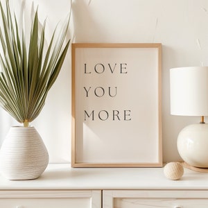 K&ouml;nnte beinhalten: Ein gerahmter Druck mit dem Text "LOVE YOU MORE" in schwarzen Buchstaben auf cremefarbenem Hintergrund. Der Rahmen ist aus hellem Holz und der Druck steht auf einer wei&szlig;en Kommode mit einer Vase mit gr&uuml;nen Pflanzen und einer Lampe.