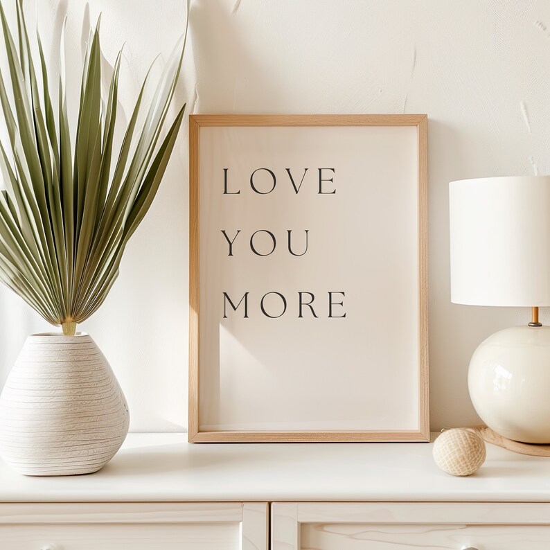 K&ouml;nnte beinhalten: Ein gerahmter Druck mit dem Text "LOVE YOU MORE" in schwarzen Buchstaben auf cremefarbenem Hintergrund. Der Rahmen ist aus hellem Holz und der Druck steht auf einer wei&szlig;en Kommode mit einer Vase mit gr&uuml;nen Pflanzen und einer Lampe.