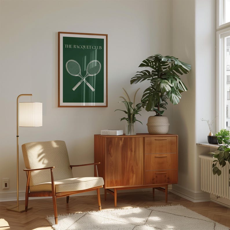 May include: A green poster with white text that reads "The Racquet Club" and a graphic of two crossed tennis rackets. The poster is hanging on a white wall in a living room with a wooden cabinet, a chair, a floor lamp, and plants.