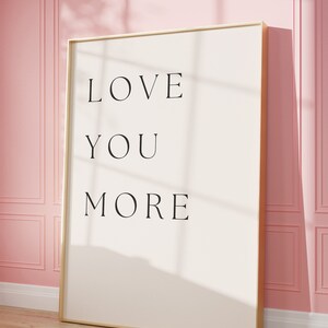 K&ouml;nnte beinhalten: Ein gerahmter Druck mit dem Text "LOVE YOU MORE" in schwarzen Buchstaben auf wei&szlig;em Hintergrund. Der Rahmen ist golden und der Druck h&auml;ngt an einer rosa Wand.