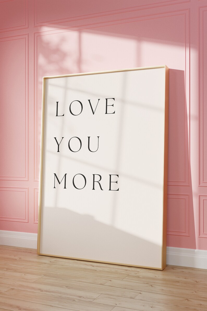 K&ouml;nnte beinhalten: Ein gerahmter Druck mit dem Text "LOVE YOU MORE" in schwarzen Buchstaben auf wei&szlig;em Hintergrund. Der Rahmen ist golden und der Druck h&auml;ngt an einer rosa Wand.